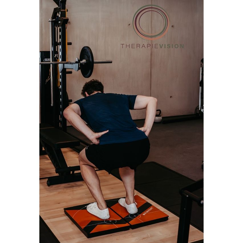 Ein Mann führt eine Übung in einem Fitnessstudio durch. Er steht auf einer Balance-Plattform, schaut nach unten und berührt seinen unteren Rücken mit beiden Händen.