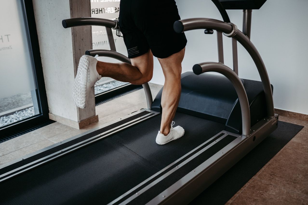Eine Person läuft auf einem Laufband in einem Fitnessstudio. Die Person trägt weiße Turnschuhe und schwarze Shorts.