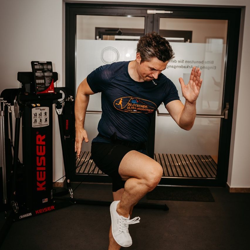 Ein Mann in einem blauen T-Shirt macht Kniebeugen in einem Fitnessstudio. Er steht mit dem Rücken zur Kamera. Hinter ihm befindet sich eine große Glastür und ein Laufband.