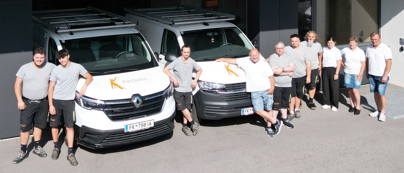Eine Gruppe von Personen steht vor zwei weißen Lieferwagen, einer mit Logo, auf einem Parkplatz unter einem Gebäude mit Balkon und Tisch.