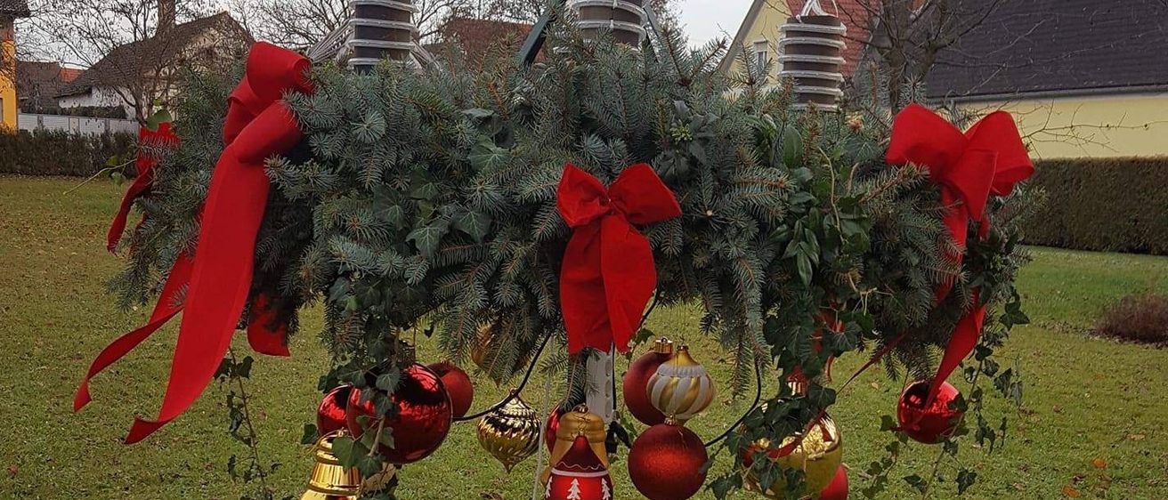 Ein Weihnachtskranz mit roten Schleifen und Ornamenten hängt an einem Metallpfahl in einem Grasfeld mit Häusern im Hintergrund.