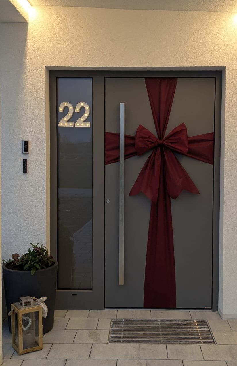 Eine Haustür mit einem roten Band und der Nummer 22 darüber. Links ein Topf mit einer Pflanze und eine schwarze Fernbedienung.