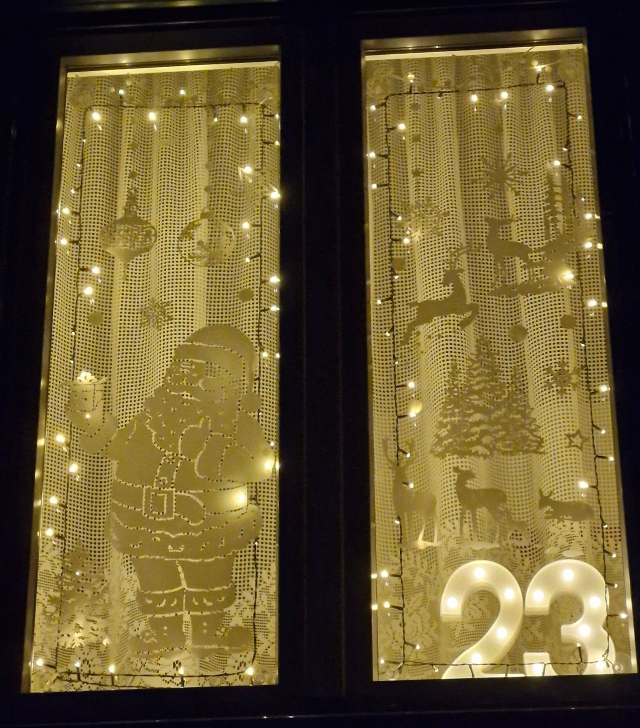 Zwei Fenster zeigen festliche Weihnachtsdekorationen, darunter eine Ausschnitt von Santa Claus mit Laterne und einer großen beleuchteten Nummer 23. Beide Fenster haben Lichterketten und Spitzenvorhänge.