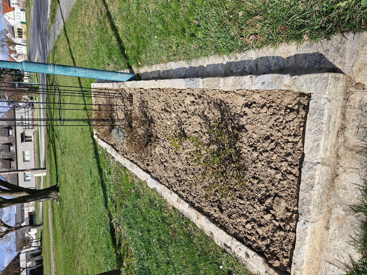 Eine Draufsicht auf ein dreieckiges Gartenbeet mit Steinumrandung und einem Metallgitter auf der linken Seite. Das Gartenbeet ist mit Erde gefüllt und hat eine kleine Pflanze darin wachsen.
