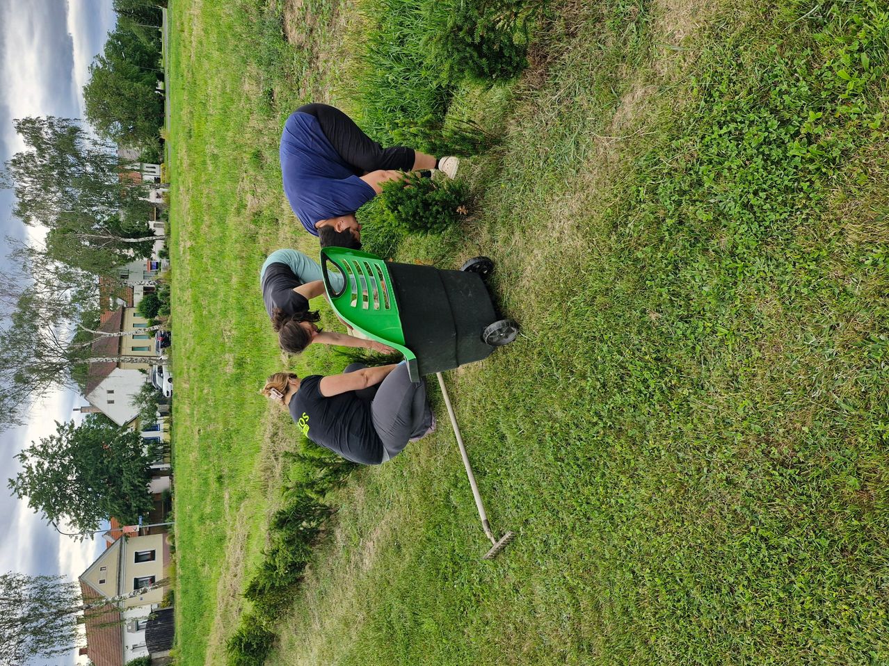 Three individuals are engaged in picking up litter in a grassy field. One is bent over, another is holding a green chair, and a third is using a rake.