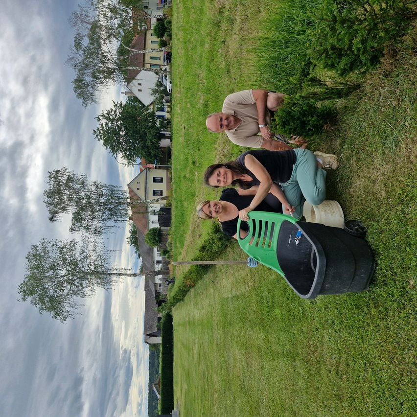 Three people are sitting in a grassy field, two women and one man. They are smiling and seem to be posing for a photo. The man is on the back, and the two women are in front.