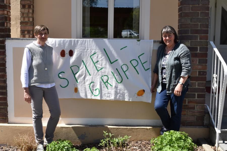Zwei Frauen stehen vor einem Backsteingebäude, eine hält ein Banner mit der Aufschrift 'SPIEL - GRUPPE'. Sie lächeln und scheinen für ein Foto zu posieren.