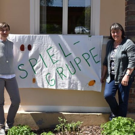 Zwei Frauen stehen vor einem Backsteingebäude, eine hält ein Banner mit der Aufschrift 'SPIEL - GRUPPE'. Sie lächeln und scheinen für ein Foto zu posieren.