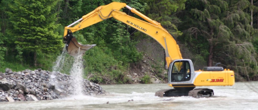 Ein gelber New Holland-Bagger schüttet Steine in einen Fluss und verursacht Wasserspritzer. Bäume und ein Flussufer sind im Hintergrund.