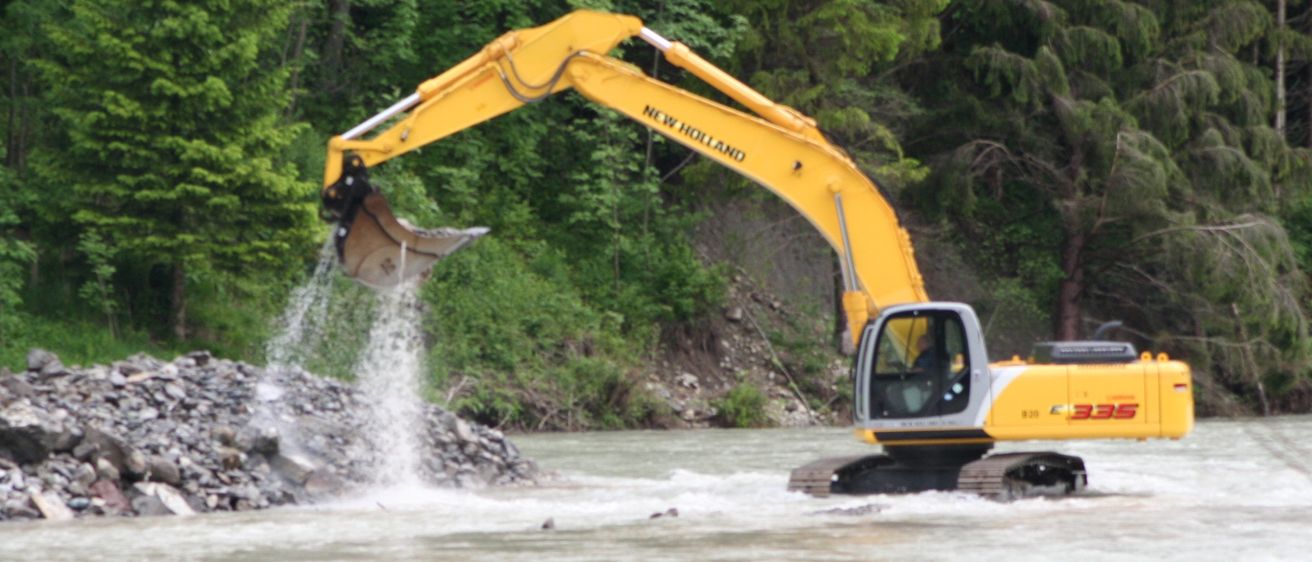Ein gelber New Holland-Bagger schüttet Steine in einen Fluss und verursacht Wasserspritzer. Bäume und ein Flussufer sind im Hintergrund.