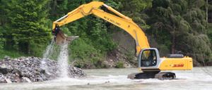 Ein gelber New Holland-Bagger schüttet Steine in einen Fluss und verursacht Wasserspritzer. Bäume und ein Flussufer sind im Hintergrund.