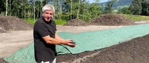 Ein Mann in einem schwarzen T-Shirt und weißen Shorts steht in einem Erdbereich mit einem grünen Planen, lächelt und hält Erde. Hinter ihm sind Haufen von Erde, und Bäume und ein Berg sind im Hintergrund.