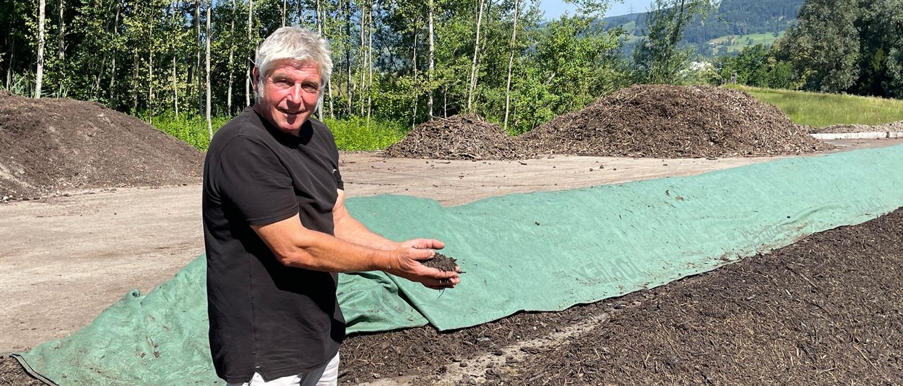 Ein Mann in einem schwarzen T-Shirt und weißen Shorts steht in einem Erdbereich mit einem grünen Planen, lächelt und hält Erde. Hinter ihm sind Haufen von Erde, und Bäume und ein Berg sind im Hintergrund.