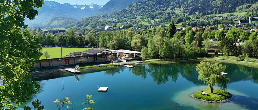 Ein ruhiger See umgeben von üppigem Grün und Bergen, mit einem kleinen Gebäude und einem Steg.