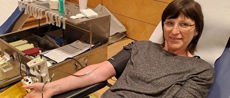 Eine Frau sitzt auf einem Stuhl mit einem Blutentnahme-Armlehne und lächelt für ein Foto. Medizinische Geräte sind in der Nähe, und eine blaue Wand mit Holzpaneelen ist hinter ihr.