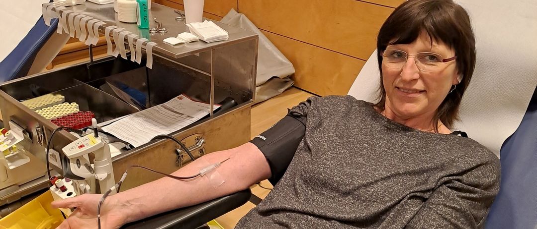 Eine Frau sitzt auf einem Stuhl mit einem Blutentnahme-Armlehne und lächelt für ein Foto. Medizinische Geräte sind in der Nähe, und eine blaue Wand mit Holzpaneelen ist hinter ihr.
