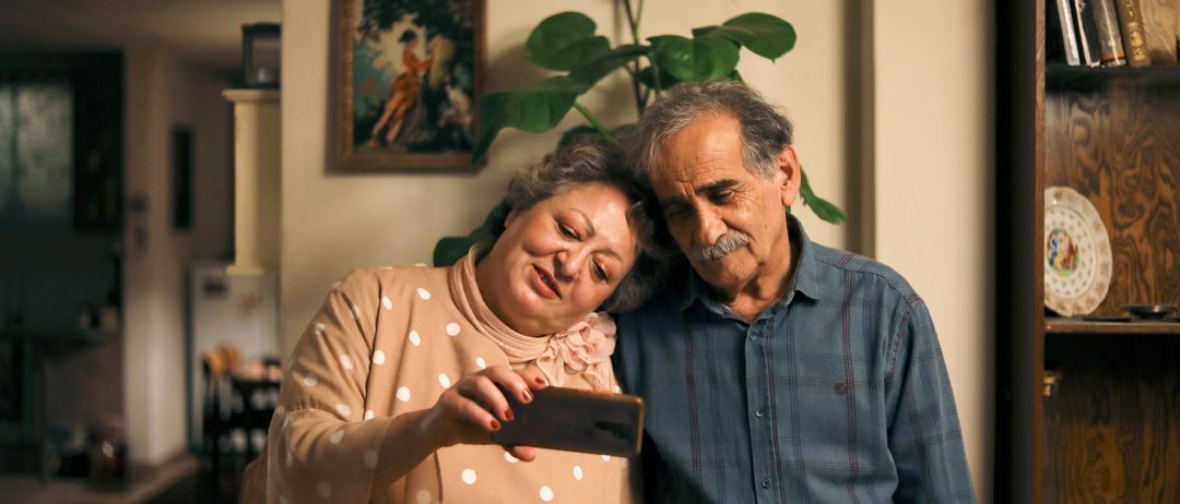 Ein älteres Paar macht ein Selfie in ihrem Zuhause, in der Nähe einer Wand mit einem Gemälde und einer Pflanze stehend.
