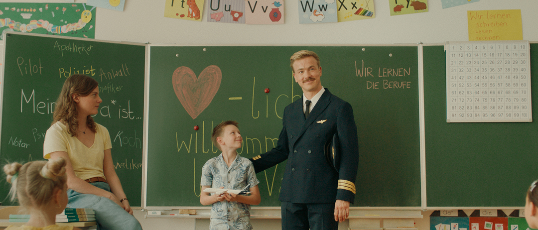 Ein Pilot steht neben einem Jungen in einem Klassenzimmer mit einer Tafel, auf der steht: 'Wir lernen die Berufe.' Der Junge hält ein Spielzeugflugzeug.