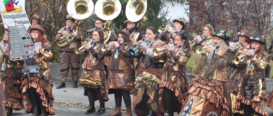 Eine Brassband in Steampunk-Kostümen spielt auf einer Straße, mit Bäumen und einem Gebäude im Hintergrund.