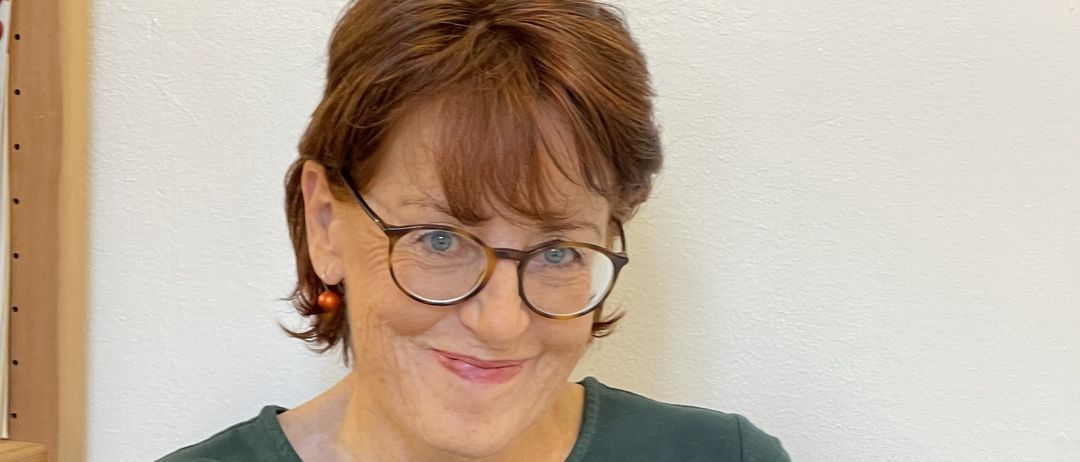 Eine Frau mit kurzen roten Haaren und einer Brille sitzt und liest ein Buch, lächelt und zeigt ihre Zähne.