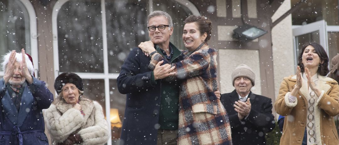 Ein Mann und eine Frau umarmen sich lächelnd während einer schneereichen Veranstaltung. Sie sind von anderen Menschen umgeben.