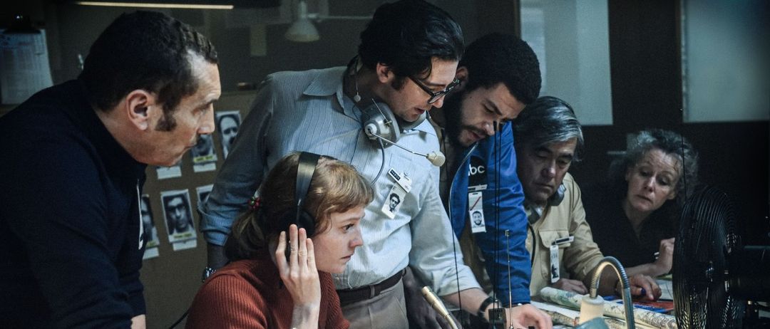 Eine Gruppe von Menschen ist um einen Tisch in einem Kontrollraum versammelt. Die Frau vor ihnen trägt Kopfhörer und wirkt konzentriert. Die anderen schauen zu ihr und scheinen zuzuhören. Sie haben Headsets auf und Mikrofone vor sich.