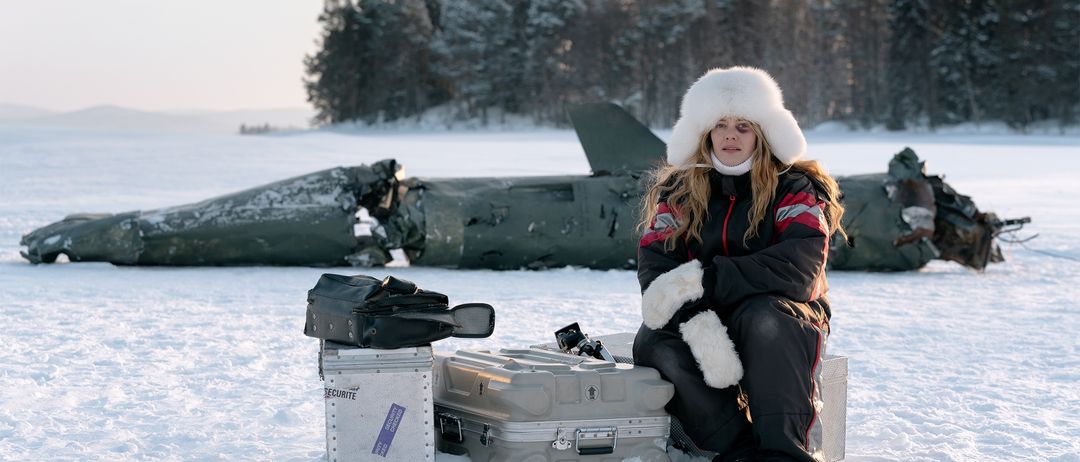 Eine Frau in einer Pelzmütze und Wintermantel sitzt im Schnee neben einem abgestürzten Flugzeug, mit Gepäck und einer Tasche in der Nähe.