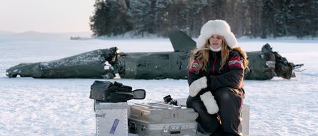 Eine Frau in einer Pelzmütze und Wintermantel sitzt im Schnee neben einem abgestürzten Flugzeug, mit Gepäck und einer Tasche in der Nähe.