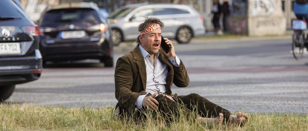 Ein Mann mit Stirnband sitzt auf dem Rasen am Straßenrand und telefoniert. Dahinter stehen zwei Personen und geparkte Autos.