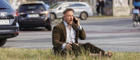 Ein Mann mit Stirnband sitzt auf dem Rasen am Straßenrand und telefoniert. Dahinter stehen zwei Personen und geparkte Autos.