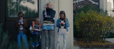 A woman and three children are standing outside, looking at their phones. The woman holds a phone in her left hand, while the children look at theirs.