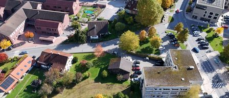 Aerial view of a town with various buildings, roads, and trees. Several cars are parked. The area is surrounded by grass and trees, with some showing autumn colors.