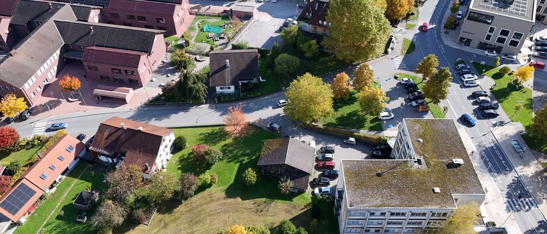 Luftaufnahme einer Stadt mit verschiedenen Gebäuden, Straßen und Bäumen. Mehrere Autos sind geparkt. Das Gebiet ist von Gras und Bäumen umgeben, einige zeigen Herbstfarben.