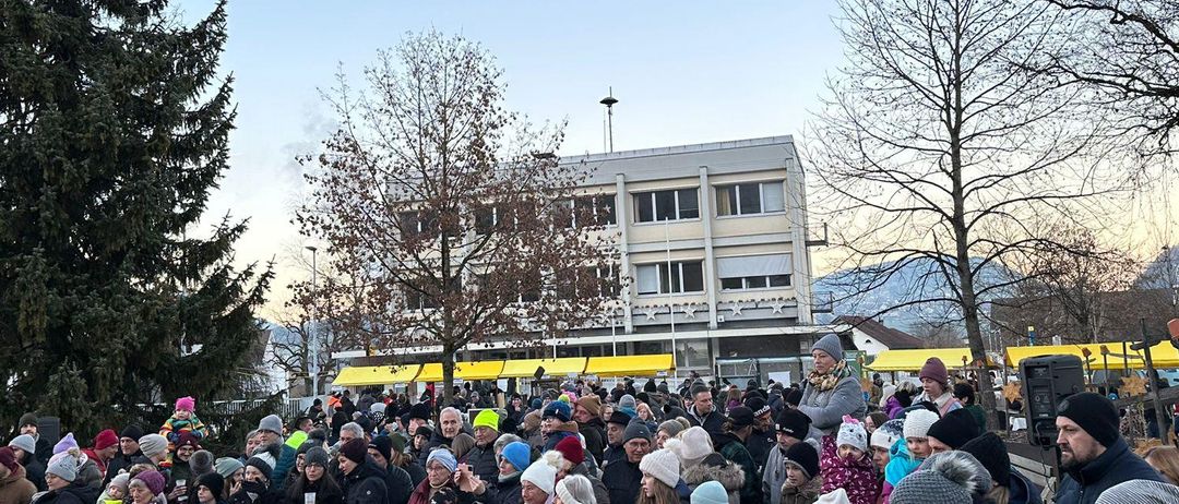 Eine große Menschenmenge, die meistens Winterkleidung trägt, ist um einen Weihnachtsbaum in einem Außenbereich versammelt. Der Bereich ist von Bäumen umgeben, und ein Gebäude mit einer Straßenlaterne ist im Hintergrund sichtbar.