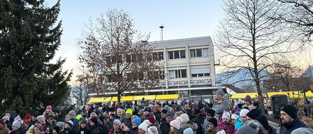 Eine große Menschenmenge, die meistens Winterkleidung trägt, ist um einen Weihnachtsbaum in einem Außenbereich versammelt. Der Bereich ist von Bäumen umgeben, und ein Gebäude mit einer Straßenlaterne ist im Hintergrund sichtbar.
