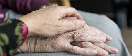 Elderly hands resting on each other with a wedding ring, one hand wearing a pink bracelet.
