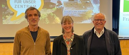 Drei Personen stehen vor einer Projektionsleinwand mit einem Gebäude im Hintergrund. Die beiden Männer tragen Jacken und die Frau ein Blumenhemd.