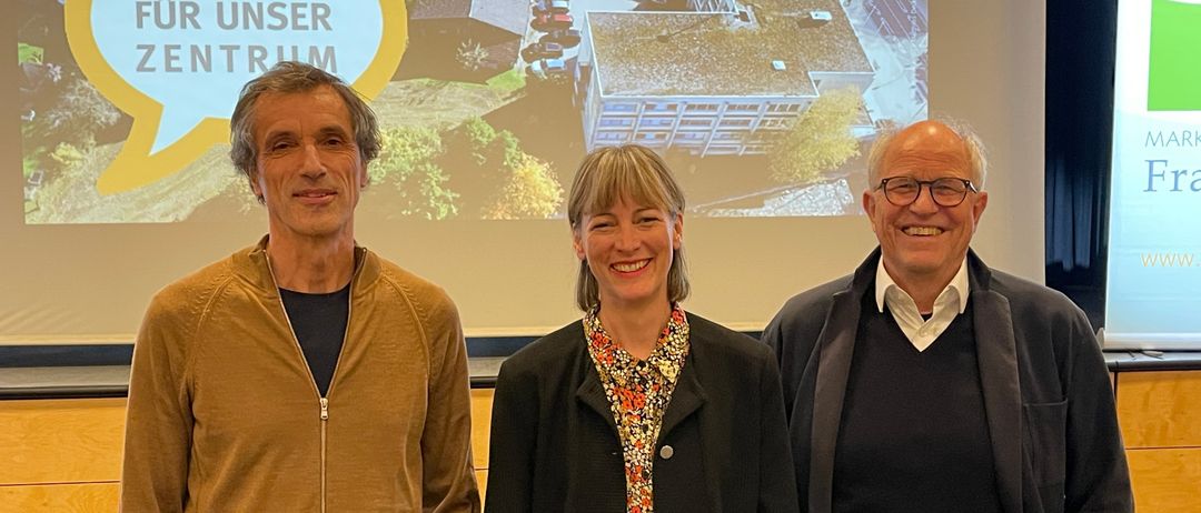 Drei Personen stehen vor einer Projektionsleinwand mit einem Gebäude im Hintergrund. Die beiden Männer tragen Jacken und die Frau ein Blumenhemd.