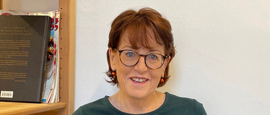 Eine Frau mit kurzen Haaren sitzt in einem Stuhl und lächelt, ein offenes Buch in beiden Händen haltend. Hinter ihr steht ein Bücherregal mit Büchern.