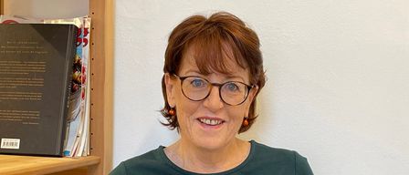 Eine Frau mit kurzen Haaren sitzt in einem Stuhl und lächelt, ein offenes Buch in beiden Händen haltend. Hinter ihr steht ein Bücherregal mit Büchern.