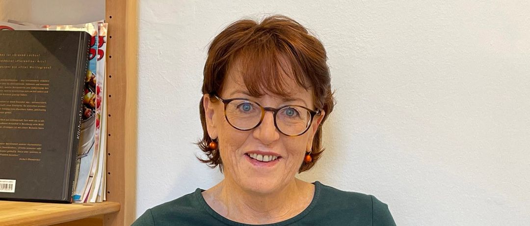 Eine Frau mit kurzen braunen Haaren und einer Brille sitzt in einem Stuhl und hält ein offenes Buch in den Händen. Sie lächelt und scheint zu lesen. Hinter ihr steht ein Holzregal mit mehreren Büchern.