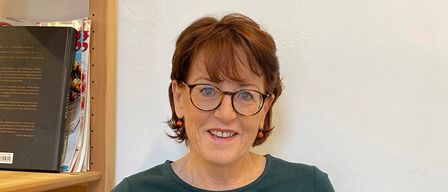 Eine Frau mit kurzen braunen Haaren und einer Brille sitzt in einem Stuhl und hält ein offenes Buch in den Händen. Sie lächelt und scheint zu lesen. Hinter ihr steht ein Holzregal mit mehreren Büchern.