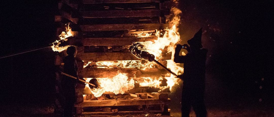 Zwei Menschen in dunkler Kleidung stehen bei Nacht neben einem Lagerfeuer, einer röstet einen Marshmallow am Stock, der andere macht ein Foto.