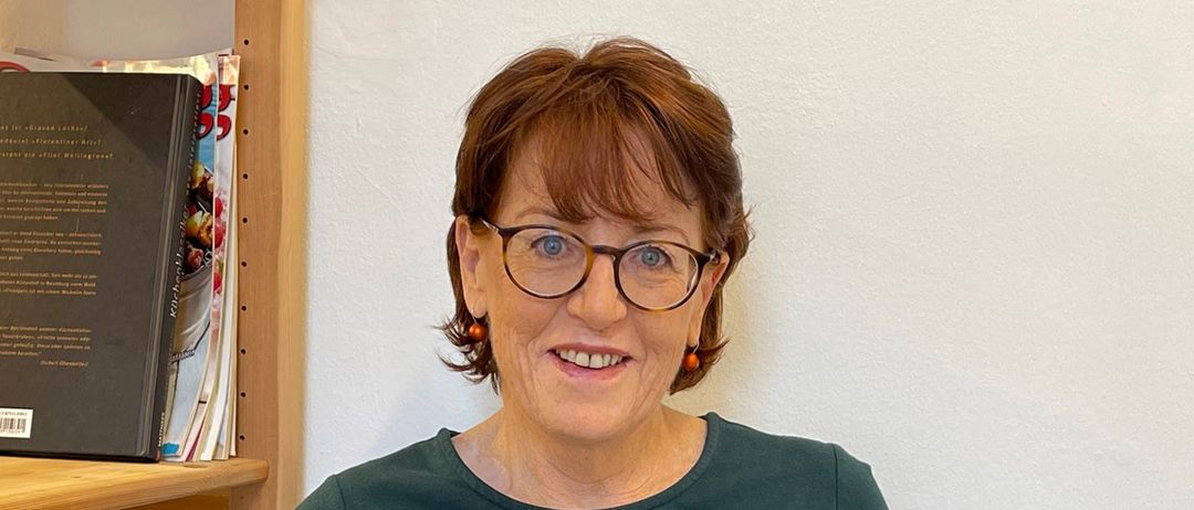 Eine Frau mit kurzen Haaren, die eine Brille trägt und ein grünes Kleid anhat, sitzt auf einem Stuhl und liest ein offenes Buch. Hinter ihr ist eine weiße Wand mit einem Holzregal, auf dem mehrere Bücher stehen.