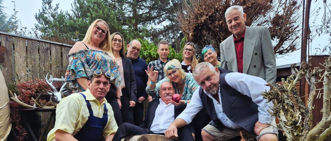 Eine Gruppe von Erwachsenen in verschiedenen Kostümen posiert für ein Foto im Garten. Sie stehen und hocken um ein Rad, einen Stein und einen Baum herum.