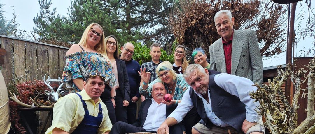 Eine Gruppe von Erwachsenen in verschiedenen Kleidungsstücken, einschließlich eines Mannes mit einem roten Apfel, posiert für ein Foto in einem Garten mit Holzstrukturen und Pflanzen.