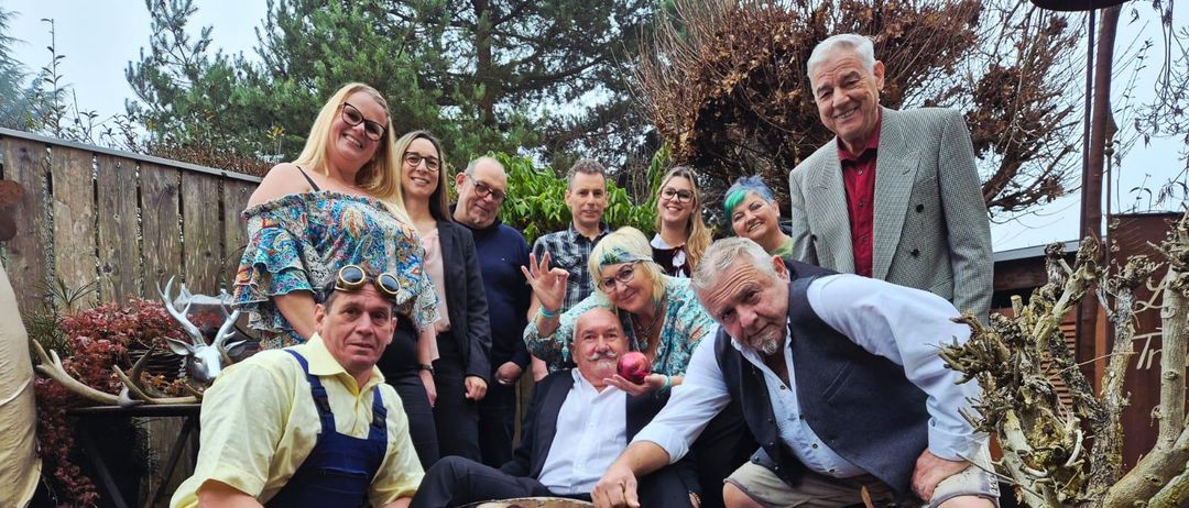 Eine Gruppe von Menschen posiert für ein Foto in einem Garten mit Holzzäunen und Bäumen. Sie sind in verschiedenen Kostümen gekleidet und einige tragen Schutzbrillen. Ein Mann hält einen Apfel.