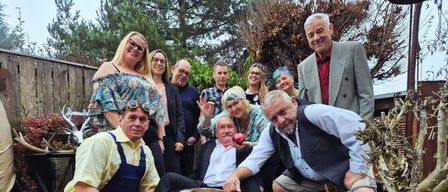 Eine Gruppe von Menschen posiert für ein Foto in einem Garten mit Holzzäunen und Bäumen. Sie sind in verschiedenen Kostümen gekleidet und einige tragen Schutzbrillen. Ein Mann hält einen Apfel.