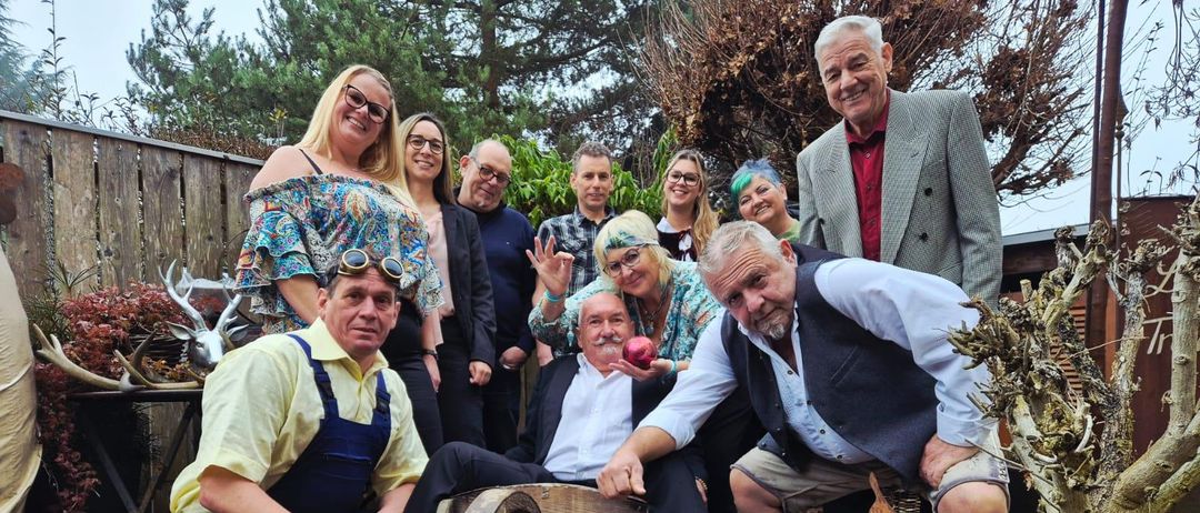 Eine Gruppe von Menschen posiert für ein Foto in einer Gartenumgebung. Einige sitzen, andere stehen dahinter. Im Vordergrund ist ein Holzrad prominent zu sehen.