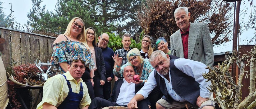 Eine Gruppe Erwachsener in verschiedenen Kostümen posiert für ein Foto in einer Gartenumgebung, einige halten Requisiten wie ein Holzrad und einen Apfel.